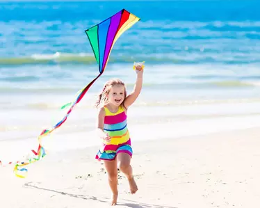 young girl running along the beach flying a rainbow diamond kite